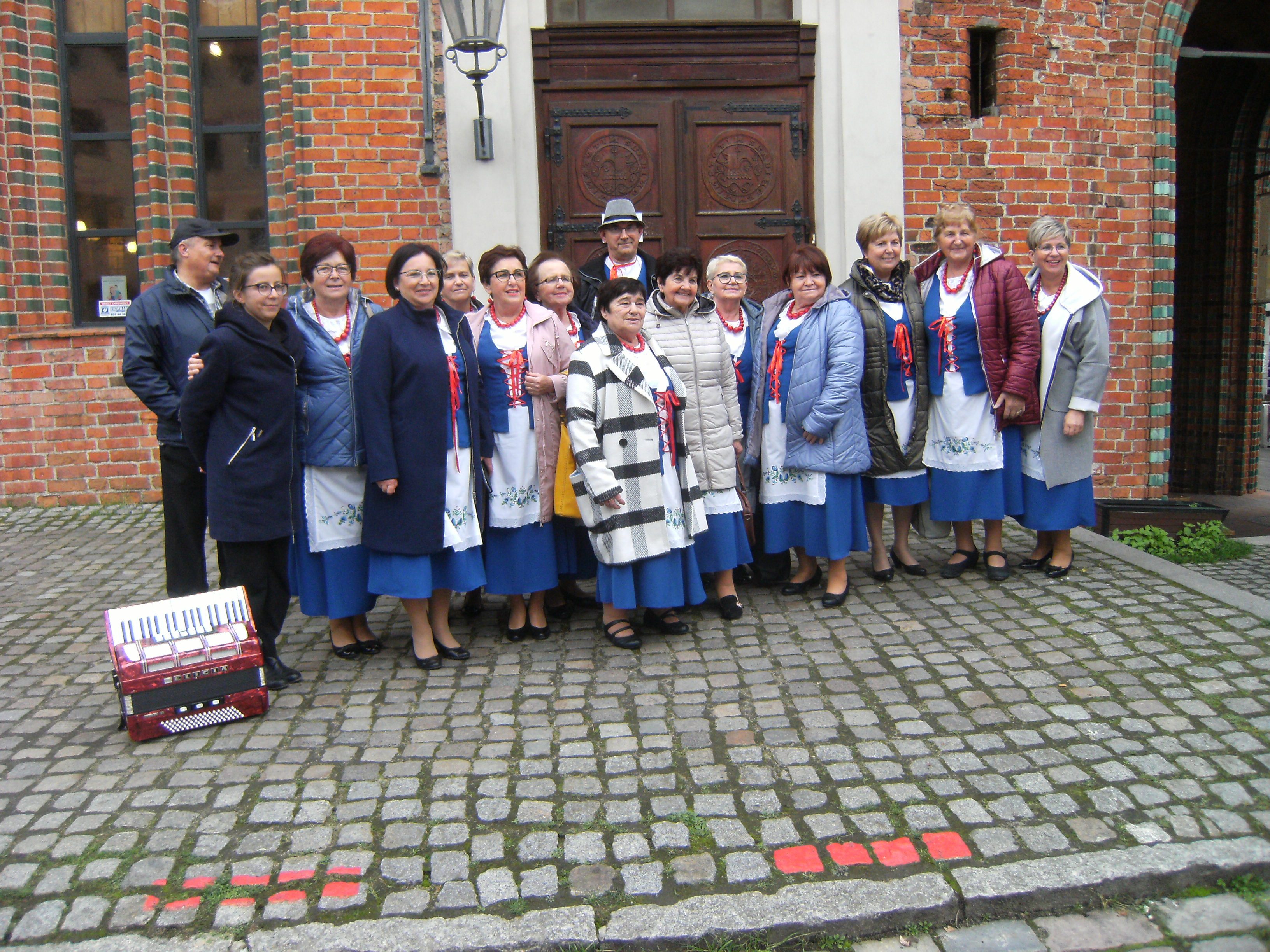 Grupa seniorów ustawiona do zdjęcia grupowego. Na tle ceglastego budynku. 